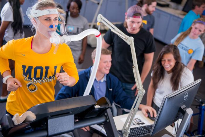 An athlete runs on a treadmill while connected to a tube measuring her breathing. Several students in the background are analyzing the data on a computer monitor.